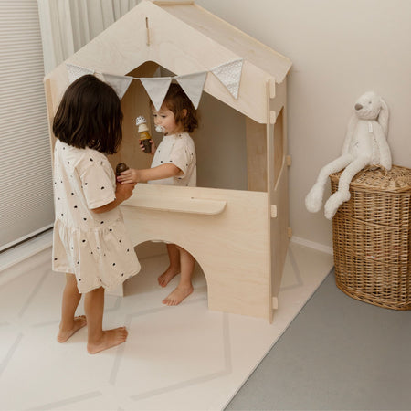 Two children playing in a wooden playhouse on the Play Mat Beni Cashmere by Eeveve, a soft and stylish EVA foam mat for indoor fun.